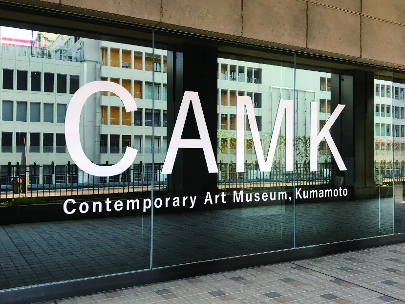 熊本市現代美術館の、街並みが映り込むガラス張りのエントランスロゴ